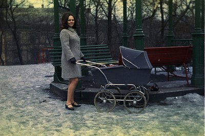 A young woman in a grey coat and dark shoes stands beside a vintage silver pram in a snowy park. Green-painted wrought iron b...