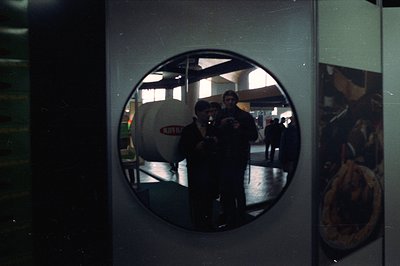 Circular mirror reflecting an interior scene. Two figures, possibly attendees, are visible within the reflection along with a...