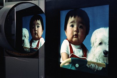 A charming, slightly grainy portrait of an infant boy in red overalls, playfully posed with a fluffy white dog. Likely a fami...