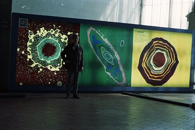 A man stands before a series of large, vibrant splash photography displays. The triptych features concentric circles of water...