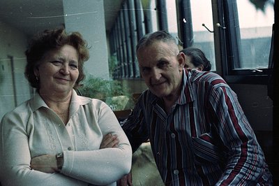 A smiling, middle-aged couple poses indoors near a window; the woman with arms crossed and the man in a patterned shirt. The ...