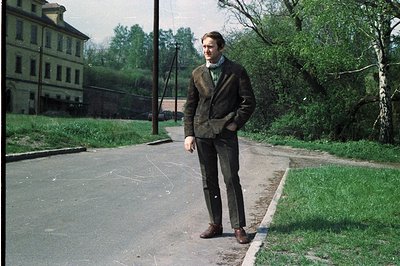 A man in a tweed suit and dress shoes stands on a roadside. A large, multi-story brick building is visible in the background....