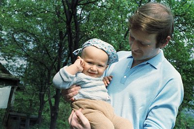 A man holds a baby wearing a patterned headband outdoors. The baby appears to be reaching out with one hand. The man is weari...