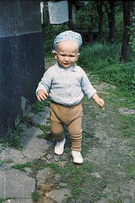 A toddler wearing corduroy pants, a light blue sweater, and a patterned headscarf stands outdoors on a cobblestone path. Gree...