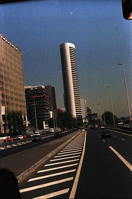 View from a vehicle on a wide, multi-lane road lined with mature trees, showcasing a dramatic skyline of modern high-rise bui...