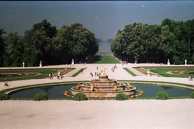 Formal garden with expansive lawns, manicured hedges, and a central fountain featuring gilded figures. The perspective emphas...