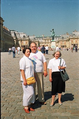 Three women pose in the courtyard of the Palace of Versailles, France. They're dressed in white and neutral-toned summer clot...