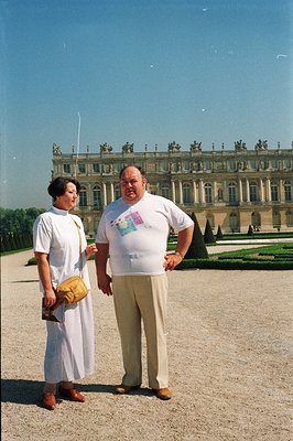 A couple poses in the formal gardens of the Palace of Versailles. The man, wearing tan slacks and a graphic tee, stands with ...