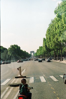 Long, straight avenue with dense tree canopy leading to the Arc de Triomphe in Paris. Heavy vehicular traffic and pedestrians...
