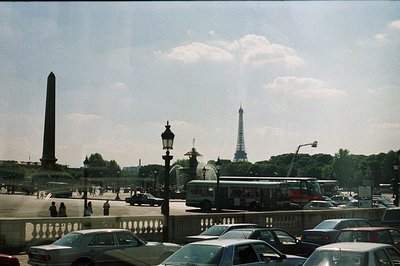 A view of the Champs de Mars in Paris, featuring the Eiffel Tower and Luxor Obelisk. A red tour bus navigates a crowded roadw...
