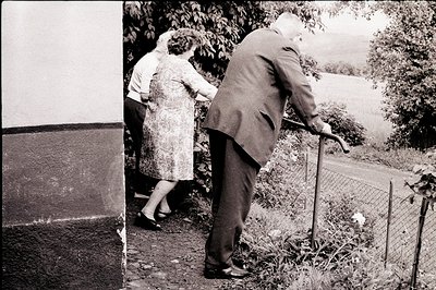 Black and white photograph depicts an elderly couple, man leaning on a cane, woman in a patterned dress, emerging from a buil...