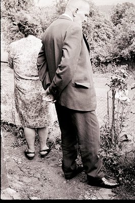 A formally-dressed man in a suit with a patterned tie stands with hands clasped behind his back. A woman in a floral dress is...