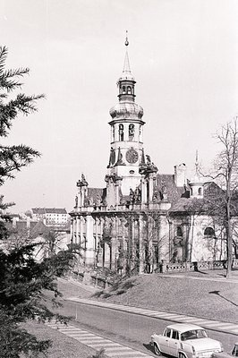 Ornate baroque church façade with a prominent tower and sculptural detailing dominates the view. A vintage car is parked alon...