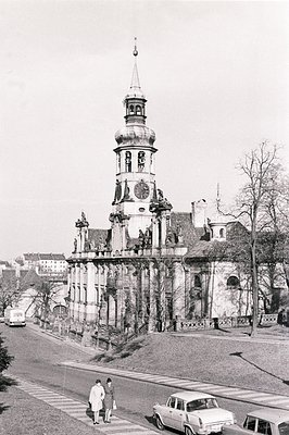 Ornate Baroque architecture dominates this view, featuring a tiered tower with clock face. Two pedestrians stroll along a str...