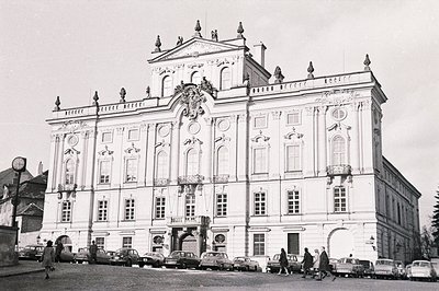 Grand Baroque palace facade with symmetrical windows & ornate detailing. Figures & vintage automobiles suggest a mid-20th cen...