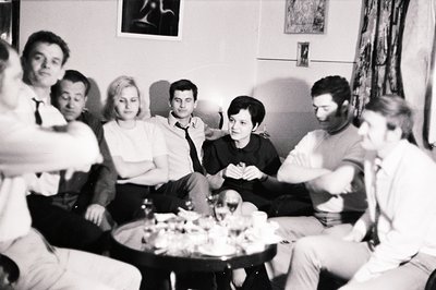 Group portrait of seven people seated on a sofa and chairs, clustered around a table laden with small bowls and glasses. Like...