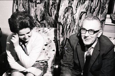 A black and white portrait shows a man and woman seated, likely in a home setting. The woman rests her cheek on her hand; the...