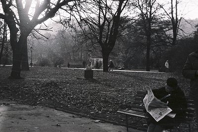 A man sits on a park bench, engrossed in reading a newspaper, amidst scattered leaves. Sparse figures populate the background...