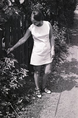 A young woman in a short, checkered dress with a patch pocket poses outdoors near a weathered wooden fence. She wears sandals...