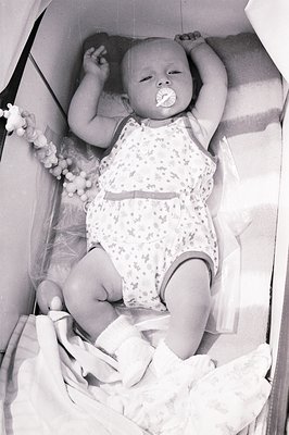 Infant, likely a baby girl, is shown lying in a crib, pacifier in mouth, wearing a patterned romper. A beaded teething toy is...