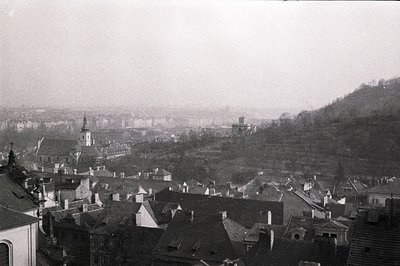 High-angle view of a European townscape with tightly-packed rooftops and a church spire. Distant buildings are visible throug...