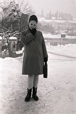 Young woman in a wool coat & knee-high boots stands on a snow-covered path, clutching a dark briefcase. A vintage phone booth...