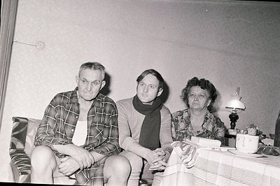Black and white portrait of three individuals seated in a domestic interior. Man in plaid shirt & undershirt; a younger perso...