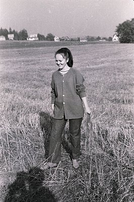 Young woman stands in tall grass, wearing a collared suit and loafers. A rural landscape, likely farmland, stretches out behi...