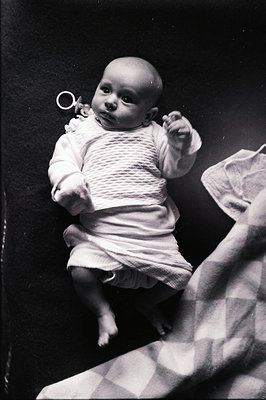 A monochrome portrait captures a baby in a textured knit romper, posed on a dark surface. The child’s gaze is direct, holding...