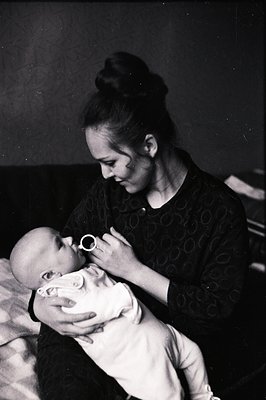 A young woman in a textured, dark dress cradles an infant, tenderly touching its face. The composition focuses on their intim...