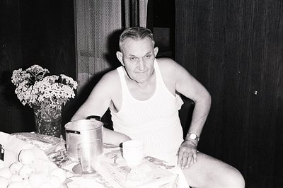 A seated, older man wearing a ribbed tank top poses in a domestic interior. He holds a wristwatch. A tabletop displays a larg...