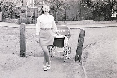 Young woman in a stylish 1960s A-line skirt suit and Mary Jane heels poses beside a vintage tricycle stroller. Architectural ...