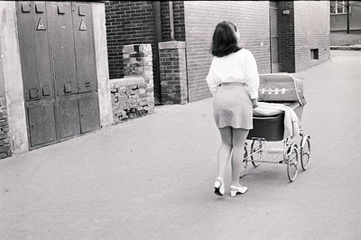 A woman walks away from the camera, pushing a vintage baby stroller along a paved street. She wears a fitted white top, a sho...