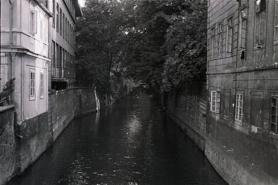 A narrow canal reflects buildings on either side. Stone architecture with varied window detailing lines the waterway. Likely ...