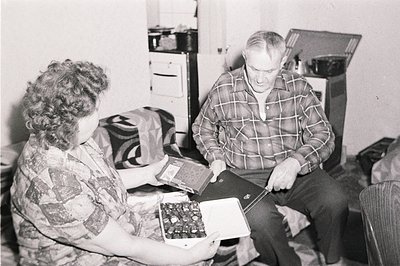 A man and woman appear to be looking at an album, with the woman presenting a box of chocolates. Interior scene, likely a hom...