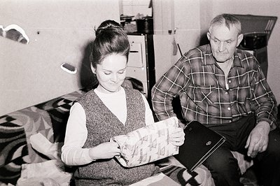 A woman with a high bun examines a wrapped gift while a man watches, seated. Interior scene reveals 1960s decor, including a ...
