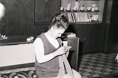 A woman with a high, textured hairstyle sits near a dark, angular cabinet, focused on sewing. The interior features geometric...