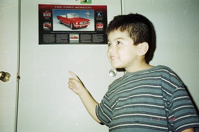 A young boy with short dark hair points upwards at a poster of a red 1964 ½ Ford Mustang. The poster is affixed to what appea...