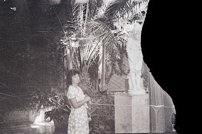 A woman in a patterned dress stands near a classical statue within a lush, possibly Mediterranean, courtyard. The scene featu...
