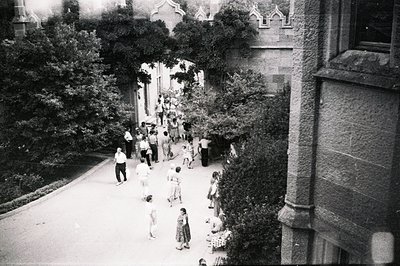 High-angle view of a lush courtyard with people strolling and children playing. Formal landscaping meets historic architectur...
