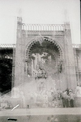 Monochrome photograph depicts a stone archway or gate, likely part of a larger structure. Group of people gathered beneath, p...