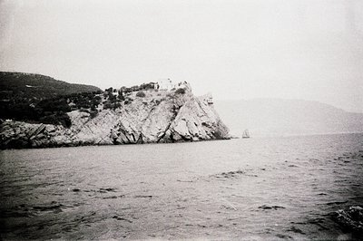Dramatic coastal scene. A rocky promontory juts into the sea, topped with what appears to be a fortified structure. Lush vege...