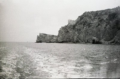 Dramatic, rocky coastline rises from a turbulent sea under an overcast sky. Evidence of human activity on the cliff top—struc...