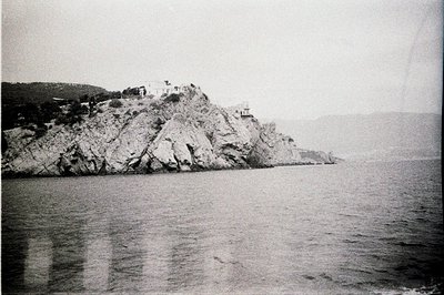 Coastal scene showing a rocky promontory topped with what appears to be a lighthouse or fortified structure. The structure is...