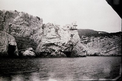 Dramatic, textured rock formations rise from a calm sea, topped by a small, architectural structure with visible windows. Lik...