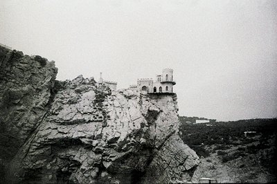 Dramatic, black and white image of a fortress or castle dramatically perched atop a rocky cliff face. Detailed stonework, arc...