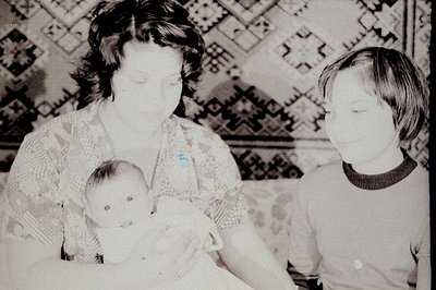 A tender moment captured: a mother cradles a baby while a young child stands nearby. The scene, set against a patterned texti...