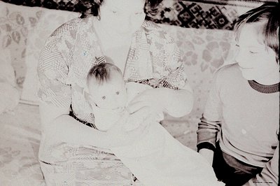 A baby is cradled in the arms of a woman, with a young boy partially visible beside them. The scene appears intimate, potenti...