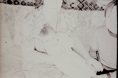 Black & white photo captures a young child, possibly an infant, nestled in a lace-trimmed cradle or bed. A woman’s arm and sh...