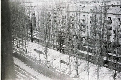 A high-angle, black-and-white image depicts a stark, post-war apartment complex courtyard covered in snow. Blocky, multi-stor...